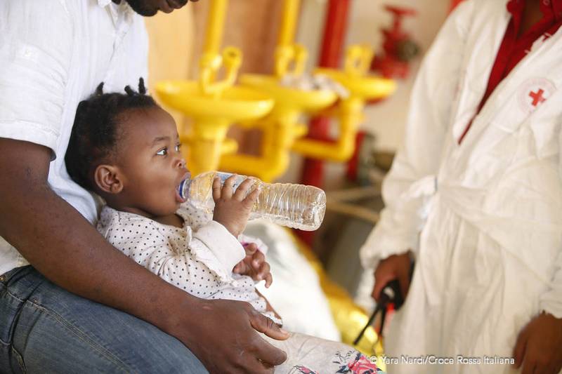 Un bimbo piccolo beve dell'acqua mentre un medico della Croce Rossa lo visita. Foto: Yara Nardi - Croce Rossa